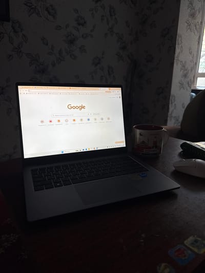 An indoor scene in Ciorescu, Moldova, displays a workspace set on a dark wooden table. A grey laptop, positioned slightly left of center, is open with its screen illuminated, showing the Google search homepage in a web browser with multiple tabs visible. To the right of the laptop, a white ceramic mug with a red interior features a colorful graphic depicting Istanbul landmarks and a red car. Further right, a white wireless computer mouse is present, alongside a dark, possibly leather, wallet or notebook. The background features dark floral wallpaper patterned with light-colored roses and vines. A window frame is partially visible on the far right, admitting diffuse natural light. The overall scene is dimly lit, with the laptop screen serving as a primary light source. No individuals are visible.