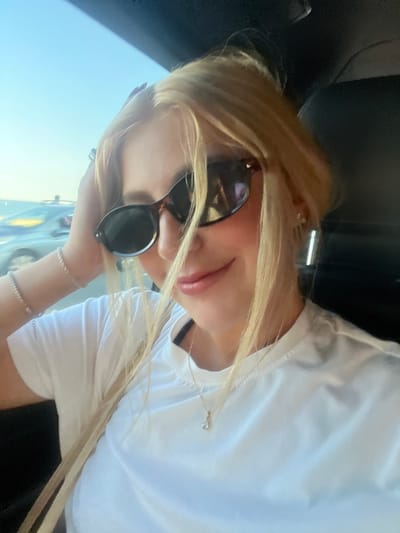 A woman with blonde hair and sunglasses is posing for a selfie. She is wearing a white shirt and jewelry on her wrist and neck. The background shows a glimpse of Cagnes-sur-Mer, France. The setting seems to be inside a car.
