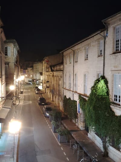 An illuminated street at night displays classic European architecture, predominantly white stone buildings with shuttered windows. Parked cars line the side of the road, while potted plants and climbing vines adorn building facades. Street lamps cast a warm glow, highlighting the narrow street's perspective and the distant figures of pedestrians and a parked van. The dark sky contrasts with the warmly lit buildings and street scene.