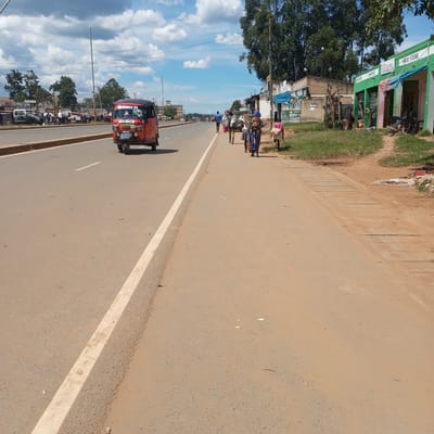 The image shows a street scene in Sheywe ward, Kenya. A red tuk-tuk is traveling on the left side of a wide paved road with a white line marking the edge. On the right side of the road, several people are walking, some carrying goods. Alongside the road are several small businesses, including a "FOOD STORE" with green and white paint and the word "AINYOZI" on its façade, as well as an electrical shop with a covered awning. A motorcycle is parked outside the "FOOD STORE". Trees are visible in the background. The sky is partly cloudy. Other vehicles and people are visible further down the road. A sign reading "HOSPITAL" and "MOKYS HIGHWAY SHOP CLINIC LANG" is seen on a building in the distance. A poster related to FIFA can be seen on the side of a building.
