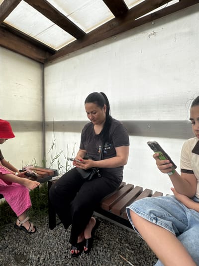 Three individuals are seated on a wooden bench in a covered outdoor area. The structure has wooden beams supporting a translucent roof, and its walls are made of corrugated plastic. The ground is covered with small gray gravel.
On the left, a young person wearing a bright pink outfit and a red bucket hat is looking down at their hands, which are holding a pair of sandals. To their right, a woman in a dark t-shirt and black pants is looking at her phone. She has her hair tied back in a ponytail and is wearing a small black purse across her body. On the far right, another young person, partially visible, is wearing a light-colored polo shirt and blue jeans, and is also looking at their phone. The overall atmosphere appears casual and relaxed, with the individuals engaged with their devices. The background plants suggest a natural or garden-like setting.