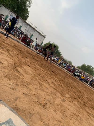 The image shows a wrestling match taking place on a sandy ground in Sokoto, Nigeria, at the coordinates 13.049 latitude and 5.238 longitude. Two men are grappling in the center of the image, while a crowd of spectators watches from the sidelines. The crowd is sitting on a mat that surrounds the wrestling area. The wrestling area is covered in brown sand. There is a white building with a green roof in the background. The sky is overcast. The wrestler on the left wears black shorts, and the wrestler on the right wears black shorts and a dark blue shirt. The wrestling match is taking place outdoors, and the background is a mix of buildings and trees. There is a man wearing a blue shirt and white shorts standing to the left of the image, in the front of the crowd. He appears to be a referee. He is watching the wrestlers. The wrestlers are all in their early 20s, and they are both wearing black shorts. They are engaged in a close struggle.