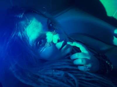 A close-up, high-angle shot features a woman lying down, her head tilted towards the viewer. The scene is illuminated by dramatic blue and green lighting, creating deep shadows and highlights. The woman has blonde dreadlocks and is wearing intricate silver face jewelry around her eyes. Her right hand is raised, with fingers touching her lips. A ring is visible on her index finger, and a bracelet adorns her wrist. The background is dark and indistinct, with shapes of light and shadow suggesting an indoor environment. The overall mood is mysterious and ethereal. No information is available about the location being in Vostochny District, Russia.