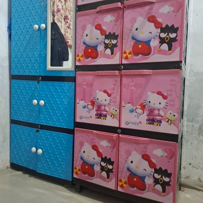 The image shows a stack of plastic storage cabinets. The left cabinet is blue with a patterned door and two white circular knobs. A mirror is affixed to the right side of the blue cabinet, reflecting a floral curtain and a dark jacket. To the right of the blue cabinet are three stacked pink cabinets. The top two pink cabinets each feature a graphic of Hello Kitty and Badtz-Maru. The bottom two pink cabinets appear to have drawers. The cabinets are positioned against a light-colored, textured wall, and the floor is a neutral-colored concrete. This scene is located in Reudeuep, Indonesia.