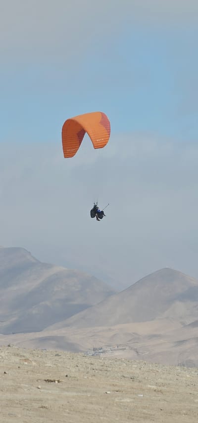 An aerial view shows a tandem paragliding flight occurring near Alto Hospicio, Chile. A paraglider with an orange canopy is in the air. Two people are visible strapped into the harness below the canopy. The environment includes a mountainous backdrop and clear blue sky. Some flat scrub and structures are visible on the ground at the foot of the mountains.