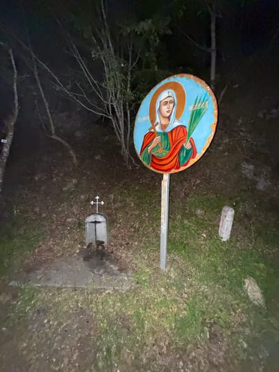 The image depicts a cemetery scene in Rila, Bulgaria. A grave marker with a Christian cross is visible in the lower center of the frame. To the left of the marker is a sign depicting an icon of a saint, possibly Saint Petka, held up by a metal pole. The ground is covered in grass, leaves, and other vegetation. A branch is seen to the left of the sign.