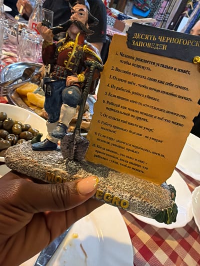A hand holds a souvenir statuette from Montenegro, inscribed with “Черногорск” in Cyrillic. The figurine depicts a man with a shovel climbing a rocky formation. Attached to the statue is a paper with "ДЕСЯТЬ ЧЕРНОГОРСК ЗАПОВЕДЕЙ" ("Ten Montenegrin Commandments") written in Cyrillic. The commandments humorously relate to avoiding work and prioritizing rest. In the background is a table set with plates and dishes, with other diners visible.