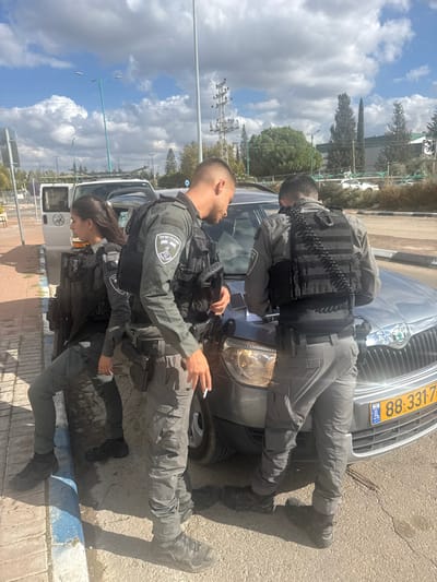 The image depicts an outdoor, daytime scene in Lod, Israel, featuring three individuals in tactical uniforms engaged with two vehicles on what appears to be a paved roadside.

A silver Skoda passenger car with an Israeli license plate (88-331-77) is positioned in the foreground, facing left. Two male personnel, dressed in olive-grey tactical pants and shirts with black tactical vests bearing Israeli Police emblems, are positioned at the front of this vehicle. The male closer to the camera has a short haircut and is looking down at the car's hood, holding a lit cigarette in his right hand. A dark rectangular object, possibly a smartphone, rests on the car's hood near his hands. The second male, with his back partially to the camera, is also focused on the car's hood, appearing to be writing or examining something; a black coiled cord extends from his tactical vest.

To the left, a female individual, similarly uniformed with grey tactical pants and a black tactical vest, leans against the open side door of a white van, which is parked behind the Skoda. Her dark hair is tied back, and she looks towards the left of the frame.

The ground consists of asphalt with a blue and white painted curb line along the left edge. The background features power lines, a street lamp, green trees, and distant structures under a partly cloudy blue sky.