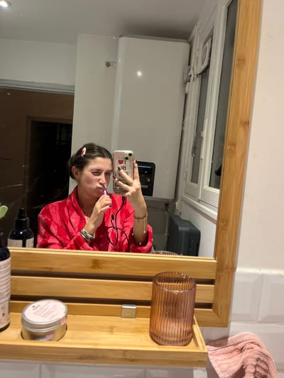 In a bathroom mirror selfie, a woman in a red robe brushes her teeth while holding a phone. Her hair is pulled back with a small clip. Reflected in the mirror are a white water heater and a window. To the right of the mirror, a wooden shelf holds a glass cup, a small tin, and various bottles. A pink towel hangs on the wall near the shelf.