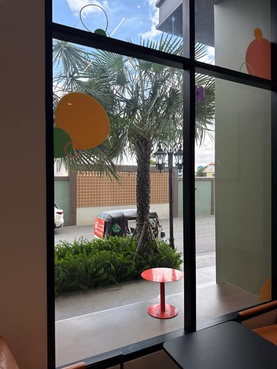 The image captures a view through a window, likely from inside a cafe or restaurant, looking out onto a street scene in Khan Mean Chey, Cambodia. The window frame is visible, and abstract decorative shapes in orange, yellow, and green are present on the glass.

Outside, a palm tree dominates the foreground, its fronds reaching towards the sky, which is partly obscured by clouds. Beyond the tree, a street with a paved sidewalk is visible. A black lamppost with two ornate lamps stands near the tree. To the right, a patch of lush green ferns adds to the foliage.

In the background, there are buildings with textured beige walls and windows. A white car is parked on the side of the street, and further down, a black vehicle with "Grab" branding and red and white signage is visible, suggesting a delivery or ride-sharing service. The presence of these elements indicates a daytime scene. The overall impression is a bright, urban environment with elements of nature interspersed.