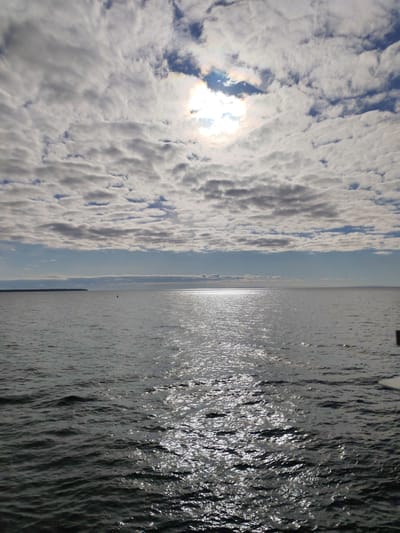 The image shows a body of water, likely a lake or sea, with a distant shoreline on the left. The water reflects sunlight, creating a shimmering effect. The sky is filled with clouds, partially obscuring the sun, which is visible as a bright spot. The overall impression is one of calm and open space. The image does not depict the city of Ozernoe Ust'e, Russia; it merely depicts an open body of water.