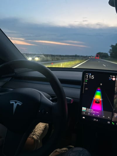 The image captures the interior perspective from the driver's seat of a Tesla vehicle, actively traveling on a multi-lane highway near Georgi-Dobrevo, Bulgaria, during the late evening or night. The time displayed on the vehicle's screen is 23:59.
The driver's left hand rests on the black, multi-functional steering wheel, which prominently features a silver Tesla emblem. The lower portion of the driver's legs and feet are visible in the lower left, clothed in dark trousers and light-colored, lace-up sneakers.
A large, vertically oriented central touchscreen display is visible to the right of the steering wheel. This screen indicates the vehicle is in "Drive" (PRND) mode, traveling at 115 km/h, with headlights activated. The battery charge is at 72%, with an estimated range of 115 units (maximum 140). The display shows a real-time visualization of the vehicle on the road, where lane lines are rendered in a rainbow gradient, indicating the activation of an advanced driver-assistance system. A depiction of a black vehicle is shown ahead of the driver's car on the display. Below this, a media player is active, displaying "MLADO GAZE" by "MOMO RECORDS, Krisko," along with playback controls. The cabin temperature is set to 21.5 degrees Celsius.
Outside the vehicle, a multi-lane asphalt highway is visible, stretching into the distance. The roadway is illuminated by the car's headlights, highlighting white lane markings and the green vegetation of the median. The horizon reveals a twilight sky with a faint band of orange and pink hues transitioning into a dark, overcast sky. A red truck with illuminated tail lights is visible in the distance on the right lane, and the headlights of an oncoming vehicle are faintly discernible further ahead on the left. The vehicle's windshield displays minor smudges or reflections.