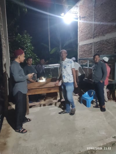 The image captures a nighttime gathering of several men in what appears to be an outdoor or semi-open space. The scene is illuminated by a bright overhead light source, creating distinct shadows on the ground.

In the foreground on the left, a man wearing a dark grey long-sleeved shirt, black pants, and a red peci (cap) stands, facing towards the right. His right hand is slightly raised, suggesting he might be speaking or gesturing. He is wearing sandals.

To his right and slightly behind him, another man in a dark shirt is partially visible, appearing to be part of the conversation. Further back, another man in a patterned shirt is seen behind a wooden structure.

In the center-right foreground, a man wearing a white short-sleeved shirt with a floral pattern, blue jeans, and sandals stands, looking towards the left. He is positioned near a wooden table or bench that occupies the central part of the image, between several of the men.

To the right of the man in the floral shirt, another man in a dark grey shirt and dark pants is visible, smiling and looking towards the left. He is also wearing sandals. Behind him and to the far right, a man in a white cap and a reddish shirt is partially visible, along with another person further back who is also wearing a white cap.

A blue plastic chair is on the ground to the right of the wooden table. The background on the left features dark foliage, indicative of trees or bushes. On the right side of the image, a brick wall structure is visible. The ground is a light-colored concrete or paved surface.

The overall atmosphere appears to be informal and social, with the men gathered and engaged in conversation.

A date and time stamp "17/02/2026 21:52" is visible in the bottom right corner of the image.