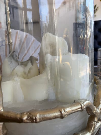 The image shows a close-up of a large, melted white candle inside a clear glass hurricane lamp. The candle has a rough, uneven surface with areas of darker discoloration, possibly from being burned. The hurricane lamp has a gold-colored bamboo-style decorative base and is reflected in the glass. In the background, through the glass, there are reflections of buildings and windows, suggesting an urban environment. The lighting is bright, with some reflections and glare on the glass. The overall impression is of a decorative candleholder. The image does not provide any information about the city of Torquay, United Kingdom, or any specific location.