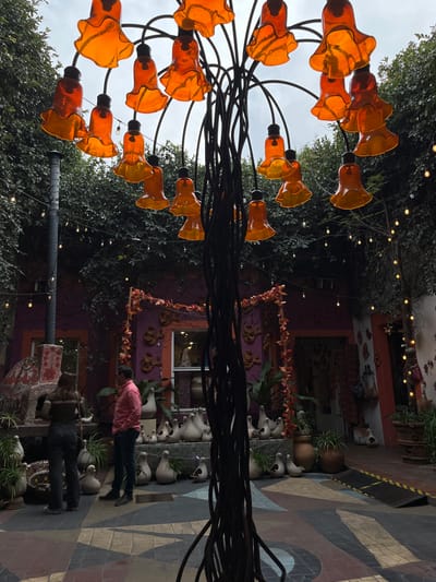 A courtyard scene featuring an ornate, wrought-iron light fixture with orange, flower-shaped shades. Two individuals stand in the walkway. One wears a pink shirt, the other dark clothing. Various potted plants line the path. A building in the background is decorated with decorative elements. Overhead, string lights are visible through the surrounding greenery. The ground is covered with a geometric tile pattern.