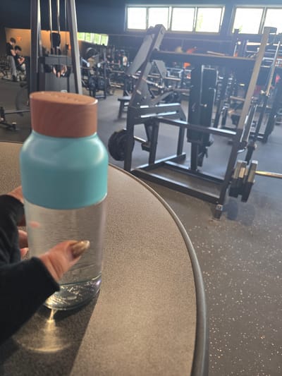 The image shows an interior view of a gym, possibly in Zirndorf, Germany. In the foreground, a person's hand is visible holding a water bottle with a brown lid and turquoise middle section. The bottle appears to be partially filled with liquid. The hand is wearing a black long-sleeved garment. The bottle is positioned over a round, speckled grey table. The gym equipment includes weight machines, a squat rack, and various other fitness apparatus. Windows on the left side of the frame provide natural light. In the background, there are other people, possibly working out. The gym floor is grey and appears to be a non-slip surface.