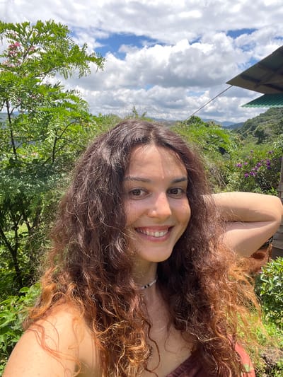In a medium close-up selfie taken outdoors, a young woman with long, wavy brown hair smiles at the camera. She is wearing a brown top and a beaded necklace. Her right arm is bent behind her head, and her left arm is not fully visible. The background features lush green foliage, including a tree with pink flowers on the left, and rolling hills with a structure in the distance. The sky above is a bright blue with scattered white clouds. The scene suggests a natural, possibly rural setting. This location is identified as Sơn Điền, Vietnam.