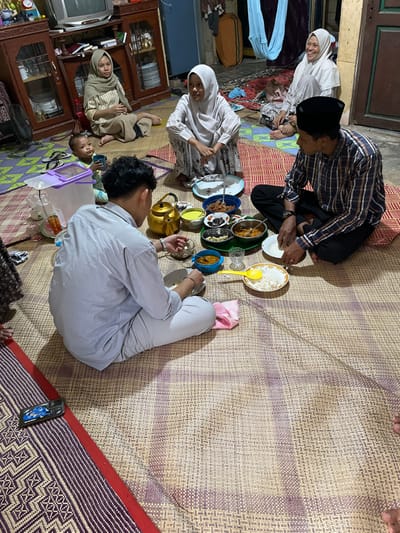 An indoor scene in Payakambuk, Indonesia, depicts six individuals, varying in age from an infant to adults, gathered for a meal. All individuals are seated directly on woven mats covering the floor. In the foreground, a male adult in a light-colored striped shirt and light pants sits cross-legged, holding a small bowl and using a spoon. To his right, an older male adult wearing a plaid shirt and a black peci (cap) sits with his legs crossed, eating white rice from a plate with his right hand. Between these two adults, an assortment of small bowls filled with various food items, including curries and side dishes, along with a golden kettle and a transparent pitcher, are arranged on the mat. To the left of the older male, a young female adult in a white hijab and patterned dress sits, facing the food. Behind her, an older female adult in a white hijab is smiling. Further to the left and back, another young female adult in a tan hijab and striped top sits observing the scene. A baby or toddler in a light blue outfit sits on the mat to the left of the foreground male, holding a small cup. In the background, wooden cabinets, a television set, and a floor fan are visible. A smartphone rests on a patterned mat in the lower left corner. The environment appears to be a residential space, characterized by modest furnishings and overhead artificial lighting.