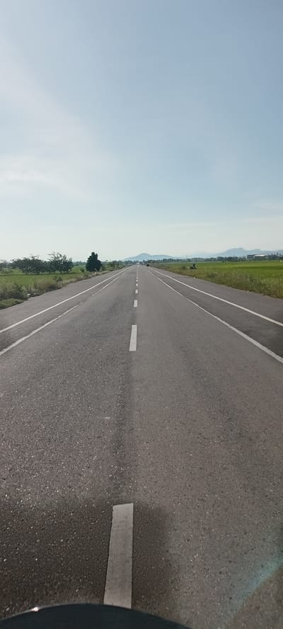 The image shows a long, straight asphalt road stretching into the distance under a pale blue sky, likely near Kutabaroh, Indonesia. The road is marked with solid white lines along the edges and intermittent white dash lines down the center. Green fields and trees border the road on either side. Distant hills or mountains are visible in the background. A vehicle is in the road in the distance. A motorcycle is parked on the right side of the road in the foreground.