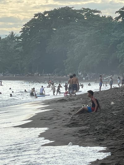 This image depicts a crowded beach in Dauin, Philippines. The foreground features dark, wet sand with white foamy waves lapping at the shore. A young man in red and blue swim trunks is sitting on the sand, facing left, with a pair of flip-flops beside him. In the middle ground, numerous people are in the water, some swimming and others on surfboards. The water appears choppy with white caps. Further back, the beach is lined with many more people, some standing and others appearing to be sitting or walking. A dense, dark green forest with palm trees lines the background of the beach. The sky above is overcast with soft, diffused light, suggesting it could be dawn or dusk. The overall scene conveys a lively atmosphere of a popular beach destination.