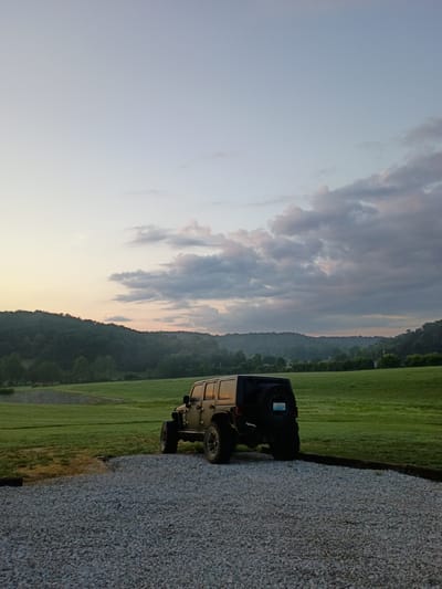 A black Jeep Wrangler is parked on a gravel driveway, facing away from the camera and slightly to the right. The vehicle is positioned in the foreground, with its rear spare tire visible. The background consists of a vast, gently rolling green field that extends towards a tree-covered hillside. The sky above is a soft gradient of light blue and pale orange, with scattered, wispy clouds, suggesting either sunrise or sunset. The scene is set in a rural environment, likely in or around Jeffersonville, United States of America. No people are present.