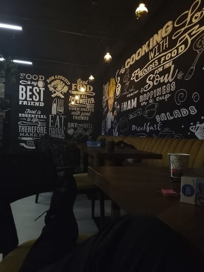 This media file captures a dimly lit, cozy interior of a cafe or restaurant in Birkirkara, Malta, with a focus on a person's legs and feet resting under a table. The time of day appears to be evening or night, indicated by the dark ambient light and the illumination from hanging pendant lights.

The main subject is a person, whose legs and feet, clad in dark shoes or boots, are visible in the foreground, suggesting a relaxed posture. They are seated at a dark wooden table, part of a booth setup with mustard yellow cushioned seating.

The most striking feature of the setting is the black wall adorned with extensive chalkboard-style white lettering and illustrations related to food and dining. Visible phrases include: "FOOD is my BEST FRIEND," "BON APPETIT," "Good FOOD is ALL THE SWEETER WHEN SHARED with good FRIENDS," "Food is ESSENTIAL to life; THEREFORE, MAKE IT GO[LD]," "EAT. DRINK and be [HAPPY/MERRY]," and "EAT WELL." Another section reads: "COOKING WITH LOVE PROVIDES FOOD FOR THE Soul," with additional words like "natural HEALTHY," "HAM HAPPINESS in a cup," "Drink Me," "SALADS," and "Breakfast." Illustrations include cutlery, cooking utensils, and various food items. A cartoon-like male chef figure is also depicted on the left side of the wall art.

On the table in the middle ground, a white paper cup with a geometric pattern, a blue pack of "Lucky Strike" cigarettes, and a pink lighter are visible. The overall atmosphere is casual and inviting, typical of a relaxed dining or social establishment.