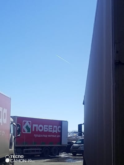 The image displays an outdoor scene in Pridorozhnyi, Russia, under a clear, bright blue sky. A single white aircraft contrail extends diagonally from the upper left to the lower right of the sky. The right side of the frame is occupied by a vertical, light-colored panel, consistent with the side of a large truck or industrial building. On the left, the front portion of a white box truck is partially visible, showing some Cyrillic text. In the midground, a prominent red box truck is parked, featuring a white 'ПОБЕДА' (Pobeda, meaning Victory) logo with a green geometric emblem, and the Cyrillic text 'продсклад честных цен' (honest price food warehouse) below it. Behind the red truck, a dark-colored van or smaller truck is partially discernible. The ground beneath the vehicles is a dark, possibly asphalt or gravel, surface exhibiting several wet patches. In the distant background, a light-colored mound, potentially snow-covered, is visible, along with sparse dark vegetation. No individuals are present in the scene. A 'TECNO CAMON 19 Neo' watermark is located in the lower left corner of the image.