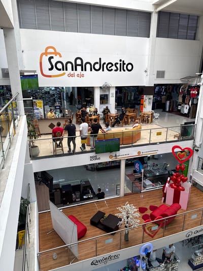 The image captures the interior of a multi-level commercial center, identified by a large sign displaying "SanAndresito del eje" and a shopping bag logo on a prominent white wall. The location is within the Area Metropolitana Centro Occidente (AMCO), Colombia.
The scene shows an open, brightly lit commercial building with white walls, large ceiling vents, and glass-and-metal railings on all visible levels.
On the uppermost visible level, a casual dining or lounge area is present, furnished with light-colored wooden tables and chairs. Approximately 10-15 individuals, mostly men in casual attire, are seated or standing in this area, some appearing to socialize. To the left, a person in a yellow shirt operates a counter, adjacent to a large screen displaying a football match. Retail clothing racks are visible in the background. Along the railing overlooking the lower levels, a group of 5-6 men are standing, while another man is seated on a curved, light-brown sofa. A smaller flat-screen television mounted on the railing also displays the football match. Below this screen, a sign reads "REDES SOCIALES" with social media handles.
The middle level features wood-patterned flooring. On the left, a red bean bag chair and a large white vertical panel stand near a dark wooden coffee table adorned with an artificial white cherry blossom tree. On the right, additional red lounge seating and several red heart-shaped floor decorations are arranged. A large, open pink and white striped gift box contains numerous red heart-shaped balloons, with three larger red heart decorations suspended above it. Retail sections with visible employees are present in the background on this level.
The lowest visible level, partially shown at the bottom, features a sign "Quintero Accesorios para el Hogar" on its fascia. This level appears to be a retail space displaying accessories and electronics, with at least two employees and a large circular ring light visible.