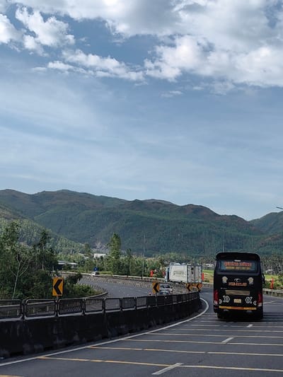 The image displays a multi-lane highway curving through a mountainous, verdant landscape under a partly cloudy sky, located in or near the city of Hòa Hiệp, Vietnam.
The upper portion of the frame shows a light blue sky with scattered white cumulus clouds. Below, a range of green, tree-covered mountains spans the horizon, with some slopes showing less dense vegetation or reddish-brown tones.
In the mid-ground, a paved highway with multiple lanes, marked by yellow dashed lines, curves sharply to the left. A dark grey concrete barrier lines the left edge of the road, featuring yellow square signs with black chevron arrows indicating the left curve. Utility poles are visible along the highway in the distance.
In the right foreground, a black passenger bus with "QUANG DUNG" and "HOTLINE: 0971.367.367" displayed on its rear window is traveling away from the viewer. The word "LIMOUSINE" is visible below its license plate, which reads "79F-001.65". Chevron-shaped decals adorn the lower rear panel of the bus. Further along the curve in the left lane, a white semi-trailer truck carrying two white shipping containers is visible, with "CONVIBER" printed on one container. Several smaller vehicles are also present on the highway. Red flags or banners are positioned along the right side of the road in the mid-ground. No individuals are discernible.