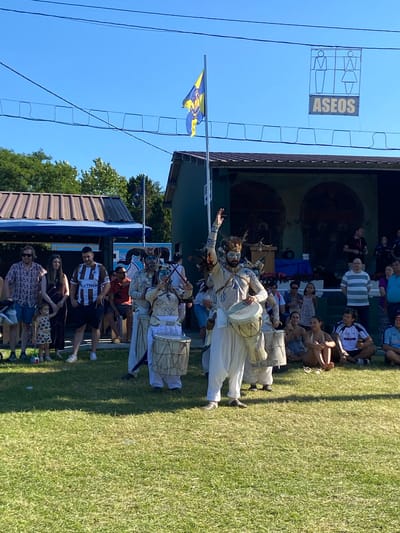 The image depicts an outdoor gathering on a sunny day, likely in the city of Llanera, Spain, given the presence of a sign that reads "ASEOS" (Spanish for "toilets"), commonly found in public spaces. In the foreground, a group of performers dressed in white, traditional-style clothing and elaborate headdresses are playing drums. One performer in the center is particularly prominent, striking a drum while raising an arm. Behind the performers, a crowd of spectators is visible, some seated and others standing. A stage or small building is situated in the background, adorned with paintings or murals. Overhead, black wires crisscross the bright blue sky, and a flag with yellow and blue elements, possibly a regional or organizational flag, is flown. The ground is covered in green grass, suggesting a park or open public area. Several individuals in the crowd are wearing casual attire, and a young child is present. The overall atmosphere appears festive and communal.