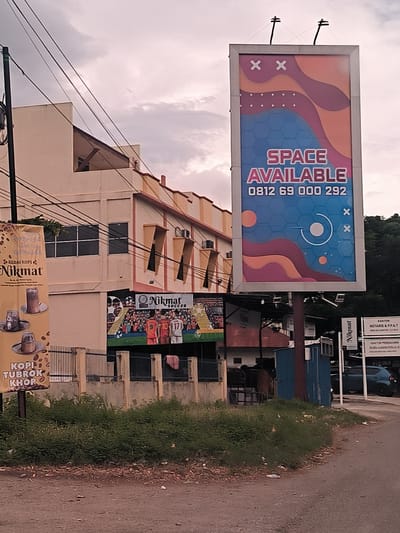 The image presents a roadside scene in Lampeuneurutgampong, Indonesia, under an overcast sky. In the foreground, a dirt path with scattered green foliage and debris leads to a paved road on the right.

The most prominent feature is a large vertical billboard on the right, displaying "SPACE AVAILABLE" in bold pink letters, along with the contact number "0812 69 000 292". The billboard's design includes abstract wavy shapes in orange and purple, set against a blue background with hexagonal patterns and decorative 'X' symbols.

To the left, a two-story building with light-colored walls and yellow accents features a unique triangular cut-out on its upper corner. Multiple power lines run across the upper left and central parts of the frame, partially obscuring the building. A yellow banner affixed to a fence in front of the building advertises "KEDAI KOPI Nikmat" and "KOPI TUBROK KHOP," showing illustrations of coffee cups. Smaller text on the banner indicates an address: "Jln. Dr. Mr. Sutan M. Hasan, Lampeuneurutgampong Aceh Besar". Below this, a mural on the building's lower section depicts football players, with visible jersey numbers 9, 10, and 17, alongside the text "Nikmat SOCCER".

Further to the right, a white sign is visible, indicating "KANTOR NOTARIS & PPAT" for "DEWI EKA SAPUTRI, S.H., M.KN." and "KANTOR PENGACARA" for "SYLANCA ZULKIFLI PUTRA, S.H., M.H.," with "WIBINA" written below. A light blue car is partially visible parked near this sign. The scene suggests a commercial area with various businesses. No human activity is directly depicted.