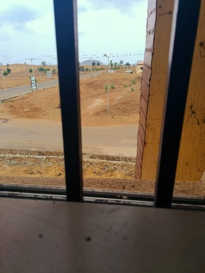 The image displays an outdoor scene in Rafin Pa, Nigeria, viewed through three dark vertical bars and a lower horizontal bar, with a textured yellow-orange wall segment visible on the right. The foreground features a paved road with double yellow lines, extending horizontally and curving to the left. Beyond this, the landscape consists of undulating, dry reddish-brown earth with sparse green vegetation, including small trees and shrubs.
Two paved roads are discernible: one in the immediate foreground and another ascending into the mid-ground. Numerous dark-poled streetlights with angled tops line these roads. Multiple white rectangular signs are present along the roads, including a stack of signs on the left side of the distant road and an additional sign further to the right. In the mid-ground on the right, a light-colored building with a dark roof is partially visible. A white pickup truck or SUV is parked facing away from the viewer on the right side of the distant road. The sky is overcast with light grey-blue clouds. No individuals, actions, or interactions are observable in the scene.
