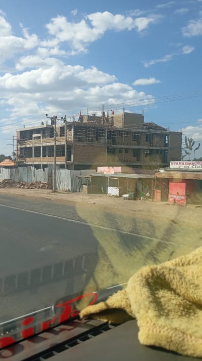 A partially constructed building, appearing to be multi-story, is the focal point, with construction workers visible on its top level. Temporary fencing surrounds the site. Several small, informal shops or stalls are situated adjacent to the construction, one marked "Nyamakima Choice Joy and Club" and another with "Nyama Choma." The scene is captured from a vehicle's interior, with a blurry dashboard and a yellow cloth partially obscuring the view. A blue sky with scattered clouds forms the backdrop.