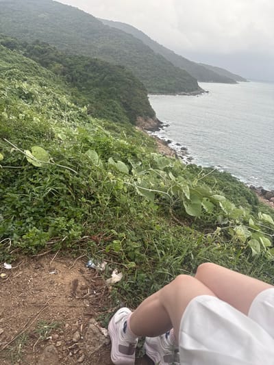The image depicts a first-person perspective from a vantage point on a vegetated hillside overlooking the coastline of Sơn Trà Ward, Vietnam. In the foreground, the legs of an individual are visible, wearing light-colored shorts or a skirt, white socks, and white athletic shoes with light purple accents. The individual appears to be seated or resting on a dirt patch littered with small pieces of trash, including plastic wrappers and paper. Immediately beyond this patch, dense green foliage, including various grasses and leafy vines, covers the steep slope leading down to the sea. The coastline features a rocky shore where the ocean water, appearing grayish-blue and slightly choppy, meets the land. In the background, multiple layers of lush green, heavily forested mountains extend along the coast and recede into the distance under an overcast or cloudy sky.