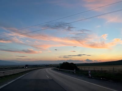 The image shows a highway at sunset, with clouds tinged pink and orange. In the distance, a truck is visible on the road, and mountains line the horizon. The scene is likely located on the highway near Churek, Bulgaria, given the presence of power lines and a rural landscape. The overall impression is one of peaceful travel at the end of the day.
