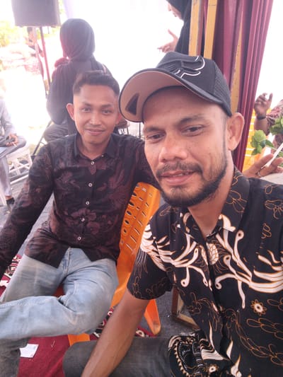 Two men are posing for a photograph outdoors. The man in the foreground, wearing a black baseball cap and a black and white patterned shirt, is looking directly at the camera with a slight smile. He has a beard and is wearing a ring on his right hand. To his left, another man in a dark, floral-patterned shirt and light blue jeans is also looking towards the camera, with a relaxed expression. He appears to be seated. The background shows glimpses of a celebratory event, with burgundy curtains and people in the distance. The scene suggests a casual gathering in a semi-urban or rural setting.