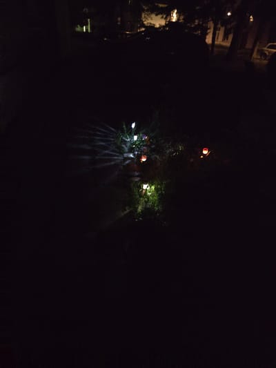 The image shows a dark outdoor scene at night, likely in a residential area of Kostelec na Hané, Czechia. In the foreground, several small, illuminated garden lights are visible, casting bright spots and rays onto the surrounding vegetation and ground. Some lights appear white, while others are colored red and orange. The lights are scattered and appear to be solar-powered decorative lights, possibly shaped like flowers or orbs. In the background, faint outlines of buildings and trees are discernible, suggesting the presence of houses or structures and natural foliage. The overall impression is of a dimly lit garden or yard at night, with artificial lighting providing the primary illumination.