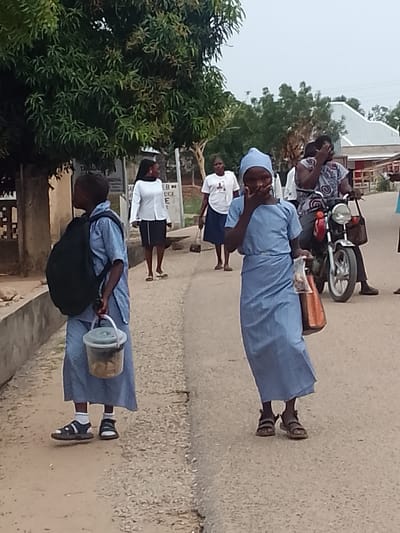 The image shows a street scene in Biliri, Nigeria. In the foreground, two girls in blue school uniforms walk on the road. The girl on the left carries a black backpack and a clear plastic bucket containing food items. The girl on the right wears a blue head covering and carries a brown plastic bag. In the background, a woman in a white blouse and black skirt walks toward the camera, while another woman in a white t-shirt and navy skirt walks nearby. To the right, a woman rides a motorcycle. A building is visible in the background on the right. A large tree is on the left side of the frame.