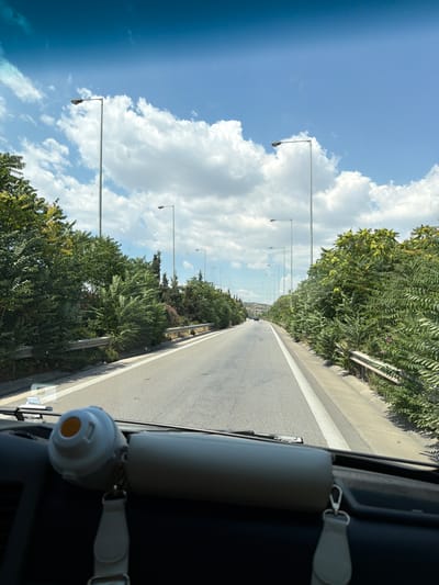 An exterior, eye-level, wide-angle shot captures a view from inside a vehicle, looking forward along a highway in Efkarpia Municipal Unit, Greece. The asphalt road, marked with a white line, curves gently to the left. On either side of the road, dense green foliage and trees line the median and the shoulders, interspersed with a metal guardrail. Tall, slender lampposts stand at intervals along the roadside, reflecting the bright, cloudy blue sky. The interior of the vehicle is visible in the foreground, with a dark dashboard and a white cylindrical object with an orange opening attached to the windshield. A grey strap hangs from the object. Distant cars are visible on the highway. The overall impression is one of travel on a sunny day through a semi-urban or suburban area.
