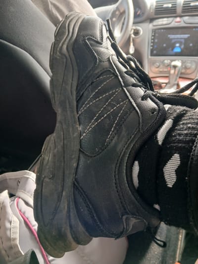 A close-up shot focuses on a person's foot wearing a black sneaker with white stitching and a black sock with a white polka dot pattern. The sneaker is slightly dirty, with visible dust on the sole and upper. The foot is resting on what appears to be car interior, with a portion of a gray fabric garment visible on the left. In the background, the dashboard of a car is visible, including the steering wheel and a digital display screen. The context suggests the person is inside a vehicle. There is no indication of the image being taken in Kuznetsovo, Russia.