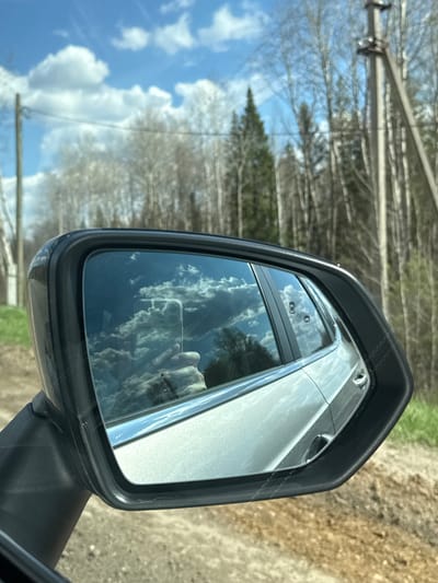 The image shows a car's side mirror reflecting a view of the sky with scattered clouds and trees. The reflection also captures the photographer's hand holding a smartphone. Outside of the mirror, a dirt road and power lines are visible. The location cannot be confirmed as Бикбарда, Russia based on the provided image.