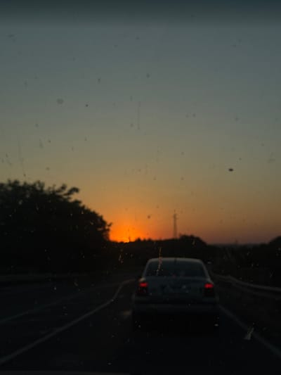 The image depicts a sunset viewed from inside a vehicle. The foreground shows a dark road with white lane markings. In the middle ground, a silver sedan is driving away from the camera, its brake lights illuminated. The background features a silhouette of trees on the left and a power line tower in the distance. The sky transitions from a pale blue at the top to a vibrant orange sunset near the horizon. The image appears to be taken in or around Belokopitovo, Bulgaria.