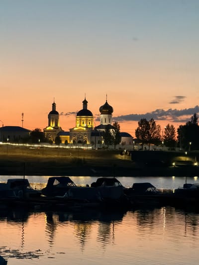 An exterior, low-angle, vertical shot captures the sunset over the Volga River in Syzran, Russia. The sky transitions from a pale blue at the top to a vibrant orange and pink gradient. In the middle ground, a brightly illuminated monastery complex stands with multiple onion domes and white facades, reflecting the warm tones of the sunset. Trees and streetlights line the riverbank, further enhancing the illuminated scene. The foreground is dominated by the dark silhouettes of several boats docked on the river, their forms softened by the dim light. The water's surface reflects the sky's colors and the lights from the monastery and streetlamps, creating a tranquil and picturesque scene. There are no people or animals visible in the frame.