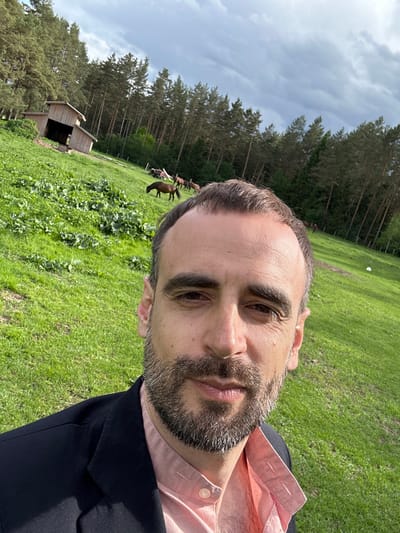 A selfie, rotated counter-clockwise, features a man with a light complexion, brown hair receding at the forehead, and a beard speckled with gray. He is wearing a pink shirt under a dark blazer. The background shows a grassy field with a small wooden structure resembling a shed and a cluster of pine trees under a cloudy sky. In the distance, several horses are visible near a fence. The location is Радашковіцкі сельскі Савет, Belarus.