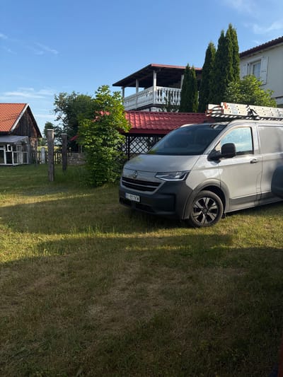 The image depicts a rural scene with a gray van parked on a grassy lawn. The van has a ladder rack on its roof and appears to be a work vehicle. In the background, there are several structures, including a wooden building with a red roof, a house with a white balcony and red trim, and several tall evergreen trees. The sky is blue with some wispy clouds, suggesting a clear day. The overall environment appears to be residential or agricultural, likely in a village setting. The license plate of the van is visible and reads "BI 811KW". There are no people visible in the image.