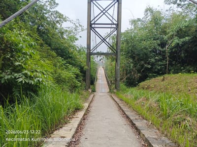 The image shows a pedestrian suspension bridge in a lush, green environment. The bridge has a concrete pathway leading up to it, flanked by tall grass on either side. A tall metal pylon, part of the suspension system, stands in the center of the frame, with cables extending from it. Dense bamboo trees and other foliage surround the bridge, creating a natural and somewhat overgrown setting. The sky is overcast. A timestamp and location are visible in the bottom left corner: "2026/06/23 12:22 Kecamatan Kuningan, Indonesia". There are no people or animals present in the image.