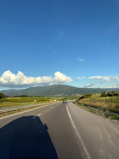 The image depicts a winding asphalt road on a sunny day, with a clear blue sky and scattered white clouds. The road curves towards the right, with a white line marking the lane. On the left side of the road, there is a grassy verge with some vegetation and a metal guardrail. In the distance, a valley unfolds, with rolling green hills dotted with small buildings, suggesting a rural or semi-rural setting. A dark-colored car is visible driving on the road, and a large shadow of an object, likely the vehicle from which the photo was taken, stretches across the asphalt. The sunlight suggests it is daytime, likely late afternoon due to the long shadow. The scene evokes a sense of travel and open space.