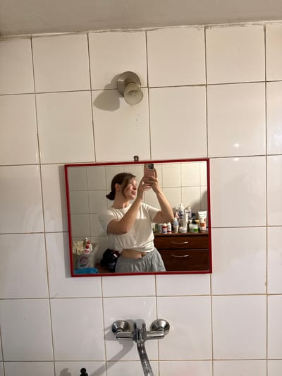 The original image, rotated 90 degrees clockwise, depicts a bathroom located in the Grcoka Urban Municipality, Serbia. The scene is primarily composed of white square wall tiles with dark, visibly worn grout lines, suggesting an older setting.
A red-framed mirror is centrally positioned, reflecting a young person taking a mirror selfie with a smartphone. The individual, with light skin and dark hair tied back, is wearing a white short-sleeved crop top and light-colored, loose-fitting bottoms. They hold a smartphone with a light-colored case up towards the mirror to capture their reflection.
Inside the mirror's reflection, behind the person, a wooden cabinet with two doors is visible. Its shelves are cluttered with numerous bottles and containers, likely toiletries or cosmetics. Below the person's reflection, on a reflected surface, are various items including a white plastic bottle, a cloth bag, a red and white container, and a blue object.
Outside the mirror's frame, to the left, a wall-mounted light fixture with a frosted bulb is affixed to the tiled wall. An adjacent, untiled wall on the far left exhibits signs of wear and dirt. To the right of the mirror, a chrome-plated faucet and tap assembly is mounted on the tiled wall. The tiles themselves show minor stains, cracks, and general wear. The time of day is not discernible, and no legible text is visible on any objects or surfaces. The person's expression appears neutral, focused on the act of taking the photo.