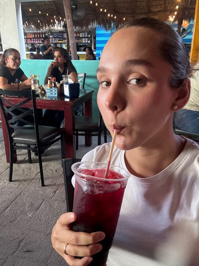 The image depicts a casual restaurant or bar in Ciudad Victoria, Mexico. In the foreground, a young adult female, with dark hair pulled back and wearing a white t-shirt, is shown from the chest up, looking directly at the camera while drinking from a large clear plastic cup. The cup contains a dark red liquid with ice and a light-colored straw positioned at her pursed lips. A gold-colored ring is visible on her left ring finger.

In the midground, to the left of the primary subject, two adult females are seated at a dark red wooden table with black metal chairs. The female closer to the camera, with dark hair, has a glass with a straw on the table. The female further back, with wavy dark hair, has a blue caddy with condiment bottles on the table in front of her.

The environment features an open-air or semi-enclosed structure with a palapa-style thatch roof from which strings of warm-toned lights are suspended. A ceiling fan is visible. A light blue and white striped wall forms part of the background. Several blurred individuals are present in the background, including one near shelves stocked with bottles, indicating a bar area. The flooring consists of grey paved stone or concrete.