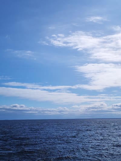 The image depicts a body of water under a sky with scattered clouds. The water is dark blue and has visible ripples. The sky is light blue with white clouds of varying sizes and shapes. The horizon line appears to be land. There are no people or other objects visible in the image. The location is difficult to determine with certainty as the image lacks distinguishing details, but it may be a lake or sea. The reference to Mologa, Russia is not confirmed.